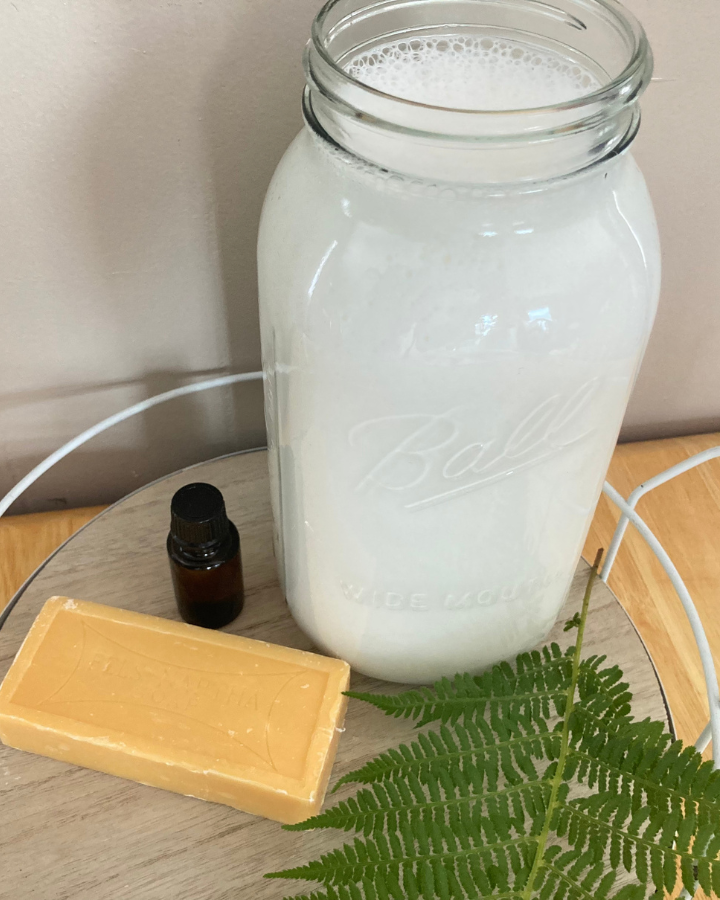 homemade laundry soap in a large mason jar with a bar of soap, essential oil and a fern leaf